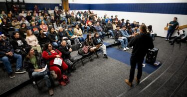Microphone at a public meeting with attendees seated and an industrial Denver backdrop beyond the windows