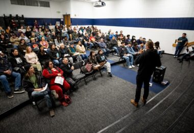 Microphone at a public meeting with attendees seated and an industrial Denver backdrop beyond the windows