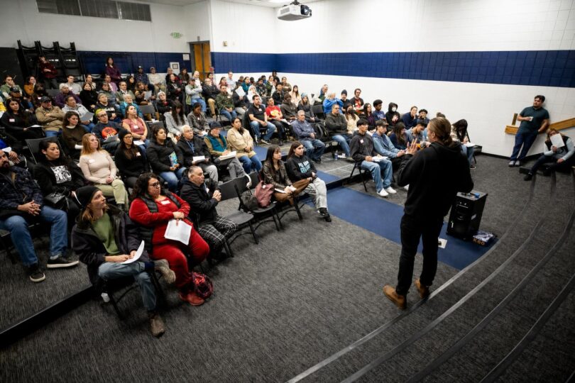 Microphone at a public meeting with attendees seated and an industrial Denver backdrop beyond the windows